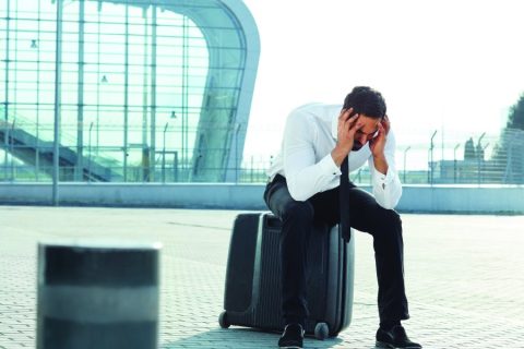A frustrated man in formal attire sits on a suitcase outside an airport terminal, holding his head in his hands. The modern glass architecture of the terminal is visible in the background, symbolizing air travel delays or disruptions. The scene conveys stress and challenges often associated with travel.