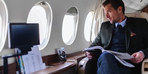 A businessman is seated comfortably in a luxurious private jet, looking out of the window while holding a document or magazine. The cabin features polished wood paneling and a built-in monitor, evoking sophistication and exclusivity during air travel.