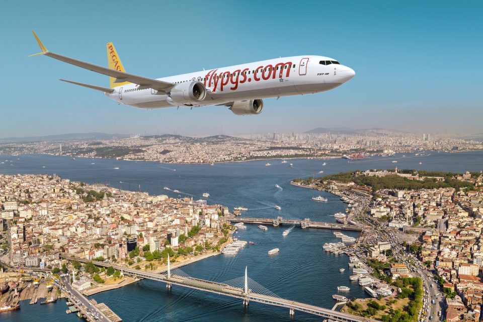 A Pegasus Airlines Boeing 737-10 aircraft soars above the scenic cityscape of Istanbul, Turkey. The plane features the airline's branding, "flypgs.com," prominently displayed on the fuselage. Below, the vibrant city spreads out with its iconic Bosphorus strait winding through, dotted with bridges, boats, and waterfront architecture. The urban skyline is framed by a clear blue sky, emphasizing the juxtaposition of modern aviation against the historic charm of Istanbul.