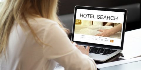 A woman seated at a desk using a laptop displaying a "HOTEL SEARCH" website interface. The screen showcases a booking portal with fields for destination, check-in and check-out dates, guests, and a search button. The image on the screen features neatly folded towels, evoking comfort and luxury. The woman, dressed in a white blouse, is actively engaged in the booking process. The environment is modern and professional, emphasizing convenience and digital efficiency in planning accommodations.
