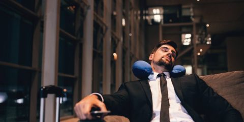 A man in a black suit with a white shirt and black tie is sitting in a comfortable chair in an airport or lounge area, resting with his head tilted to the side. He wears a blue travel neck pillow for support. The background reveals a dimly lit, modern architectural space with large glass windows and structural beams. His suitcase handle is partially visible nearby, and he looks relaxed yet exhausted, possibly reflecting travel fatigue or burnout.