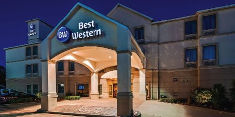 Exterior view of a Best Western hotel at night, illuminated with soft lighting highlighting the building's arch and entrance. The logo "BW Best Western" is prominently displayed above the entrance. Surrounding landscaping is visible in the foreground.