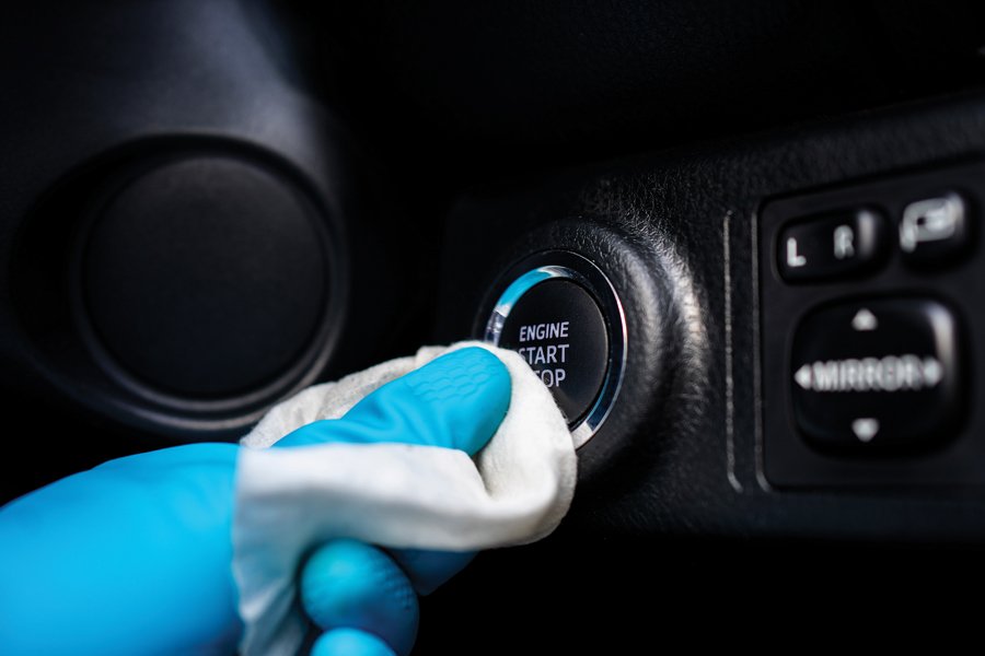 A hand wearing a blue latex glove is sanitizing a car's "Engine Start/Stop" button with a white disinfecting wipe. The close-up view emphasizes cleanliness and hygiene, focusing on vehicle interiors and safety protocols. The glove and wipe signify precautionary measures, particularly in shared or public-use vehicles.