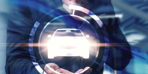 A business professional in a suit holds a glowing car icon surrounded by a futuristic circular interface, symbolizing innovation and technology in the automotive industry. The blue-toned background and light streaks emphasize a modern, high-tech atmosphere, possibly representing advancements in digital automotive services, smart vehicles, or mobility solutions. The composition highlights the integration of technology in the automotive sector.