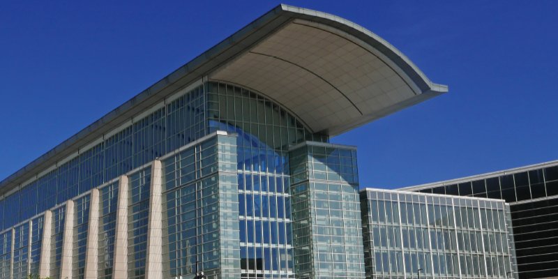 A modern architectural structure featuring a curved roof and extensive glass-panel facades, reflecting the clear blue sky. The design incorporates large vertical pillars and angular shapes, conveying a sense of contemporary elegance and functionality. This image represents a convention center or urban landmark, emphasizing innovation and architectural sophistication.