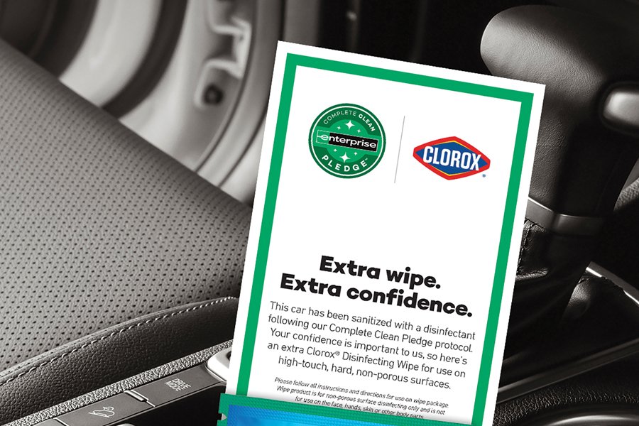 A printed notice placed inside a car, featuring logos of Enterprise and Clorox with the headline "Extra wipe. Extra confidence." The text explains the car has been sanitized according to the Complete Clean Pledge protocol and includes a complimentary Clorox Disinfecting Wipe for high-touch surfaces. The notice rests on a car seat, emphasizing cleanliness and safety in vehicle rentals.