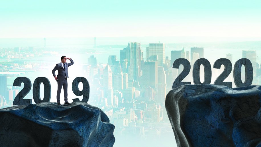 A businessman in a suit stands on a rock labeled "2019," shielding his eyes with one hand as he looks toward a distant rock labeled "2020." The two rocks are separated by a gap, representing a transition or challenge. In the background, a city skyline under a clear sky creates a sense of ambition and progress. The image symbolizes looking forward to the future, navigating change, and bridging the gap between two years.