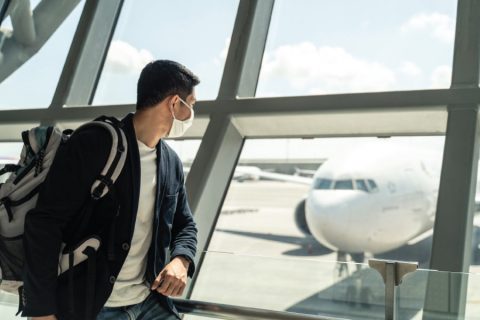 A man wearing a face mask looks out of a large glass window at an airport, observing an airplane parked on the tarmac. He carries a backpack and wears a casual blazer with a white T-shirt, reflecting a traveler preparing for a flight in the context of COVID-19 precautions. The image captures a bright, sunny day, emphasizing the modern, reflective environment of the airport terminal.