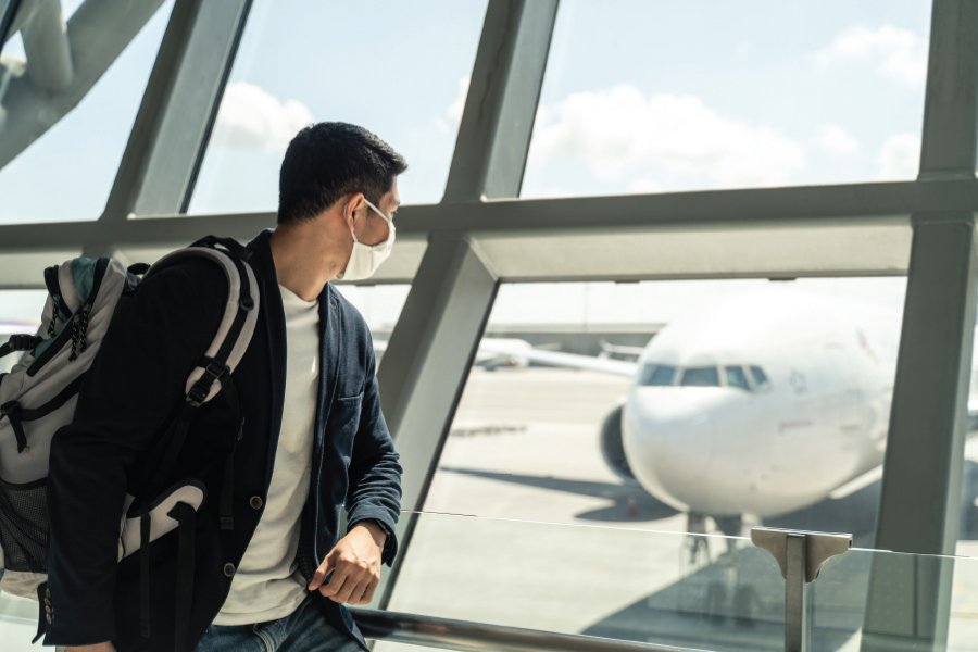 A man wearing a face mask looks out of a large glass window at an airport, observing an airplane parked on the tarmac. He carries a backpack and wears a casual blazer with a white T-shirt, reflecting a traveler preparing for a flight in the context of COVID-19 precautions. The image captures a bright, sunny day, emphasizing the modern, reflective environment of the airport terminal.