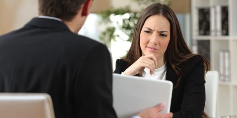 A young businesswoman sits at a desk, listening intently with a slightly perplexed expression. She is dressed in a professional suit, with her hand thoughtfully resting on her chin. Across from her, another professional, partially visible, holds a laptop and appears to be presenting or explaining something. The modern office setting in the background features bookshelves and natural light, emphasizing a professional and collaborative atmosphere.