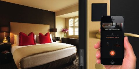 A luxurious hotel room featuring a large, neatly made bed with golden beige bedding and two vibrant red pillows. In the foreground, a hand is holding a smartphone displaying a digital room key interface. The smartphone screen shows options to unlock the door and manage the room’s smart features. The door handle and lock mechanism are modern, emphasizing advanced technology and convenience. The room is well-lit with natural light streaming in through wooden blinds, and a vase of flowers adds a touch of elegance to the decor.