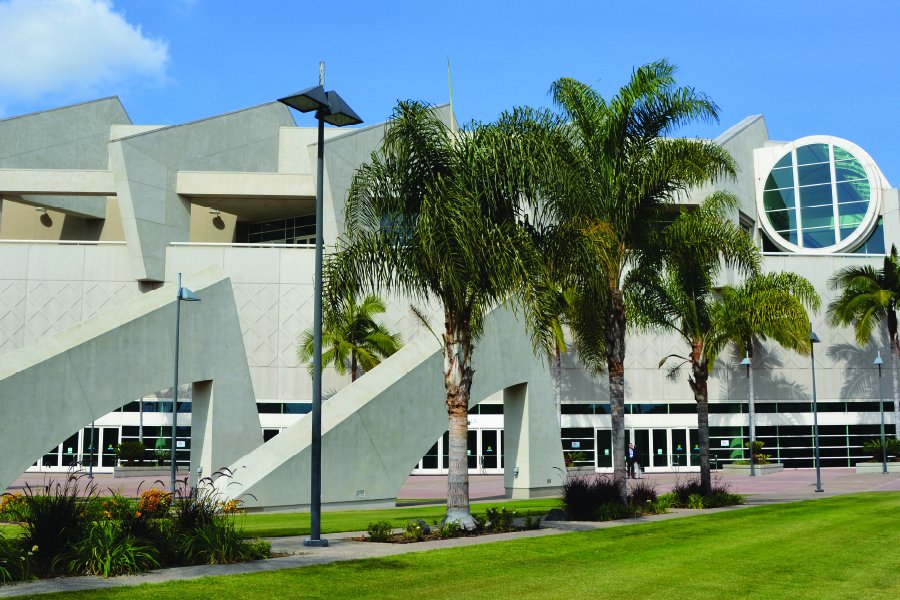 A modern architectural building with geometric designs and large windows, surrounded by palm trees and a well-maintained lawn. The structure features angular shapes and a prominent circular window, suggesting a convention center or a similar public space in a warm climate. The sky is clear, adding to the bright and inviting atmosphere.