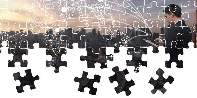 A creative composition showing a puzzle overlaying a city skyline at sunset. The pieces of the puzzle are being removed or assembled, revealing the cityscape below. Superimposed on the puzzle are white digital network connections representing global connectivity. On the right side, a professional in formal attire interacts with a tablet, blending technology and the concept of problem-solving in an urban setting.