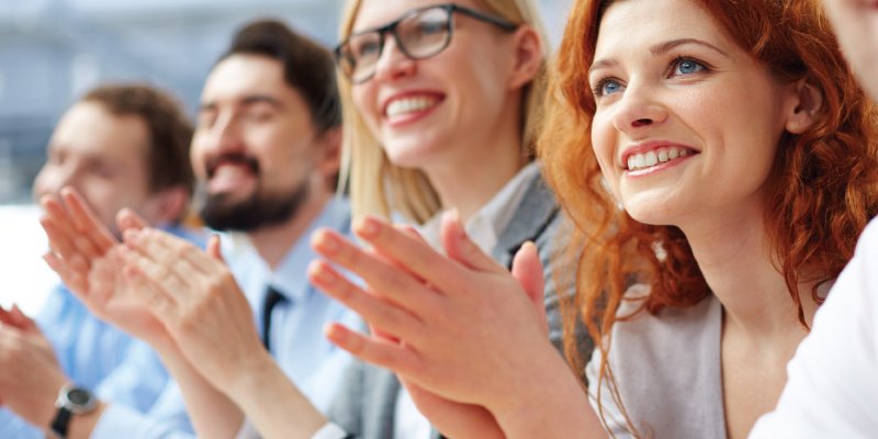 A group of diverse professionals sitting together, smiling and clapping in appreciation. The focus is on a red-haired woman in the foreground with others slightly blurred in the background, reflecting a positive and engaged atmosphere, likely at a meeting or event.
