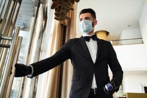 A hotel staff member dressed in formal attire, including a black suit and bow tie, wearing a face mask and gloves, holds a thermometer in one hand while opening a door with the other. The setting features a modern and elegant interior with floor-to-ceiling curtains and a bright atmosphere. The image reflects safety measures and professionalism in the hospitality industry during the pandemic.