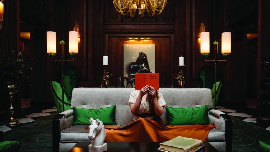 A luxurious and elegant hotel lobby with rich, dark wooden paneling and soft lighting. A person sits on a cream-colored sofa, partially hidden behind a bright orange book they are reading. The sofa is adorned with vibrant green throw pillows, matching the green accents throughout the room, including chairs and decor. A white horse sculpture and an open book rest on the coffee table in front. A large chandelier hangs above, casting a warm glow, while decorative lamps and gold details enhance the opulent ambiance. A framed artwork featuring a horse is visible in the background.