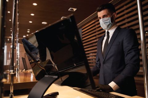 A hotel concierge or front desk staff member wearing a face mask and gloves stands behind a desk with a large computer monitor. The setting includes protective barriers and a modern, warmly lit interior design with wood paneling in the background, emphasizing safety measures and professionalism in hospitality.