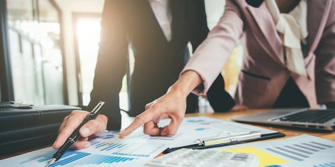 Two business professionals are engaged in a discussion, analyzing charts and graphs on a desk. One individual points to a specific area on the document, emphasizing key data, while the other holds a pen. A laptop, clipboard, and calculator are also visible, creating a focused work environment.