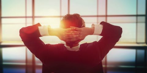 A man in a business suit sits relaxed with his hands clasped behind his head, gazing through a large window that reveals a warm, glowing sunset. The lighting creates a serene and contemplative atmosphere, emphasizing a moment of relaxation and reflection. The blurred indoor setting contrasts with the vibrant outdoor view, symbolizing a pause amidst a busy professional life.