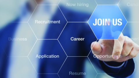 A professional recruitment-themed image with a futuristic interface. A blurred figure in business attire points to a hexagonal "JOIN US" button that is highlighted in blue. Surrounding the main button are other translucent hexagons displaying keywords such as "Recruitment," "Career," "Opportunities," "Application," and "Now Hiring." The background is softly blurred, emphasizing the interactive nature of the recruitment process.