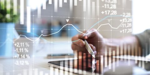 A dynamic financial scene featuring a person's hand holding a pen, interacting with a tablet. Overlaid are illuminated financial graphs and data, including percentage values and stock trends, representing analysis and decision-making in business or investment. The background is softly blurred to emphasize focus on the technology and analytics.