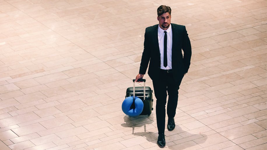 A professionally dressed man, wearing a black suit, white shirt, and black tie, walks confidently through a spacious tiled airport or transit terminal. He is pulling a small black carry-on suitcase with a bright blue travel pillow attached to the handle. The floor is made of large beige tiles, creating a neutral and polished background. The man appears focused and ready for travel, combining a sense of business and comfort.