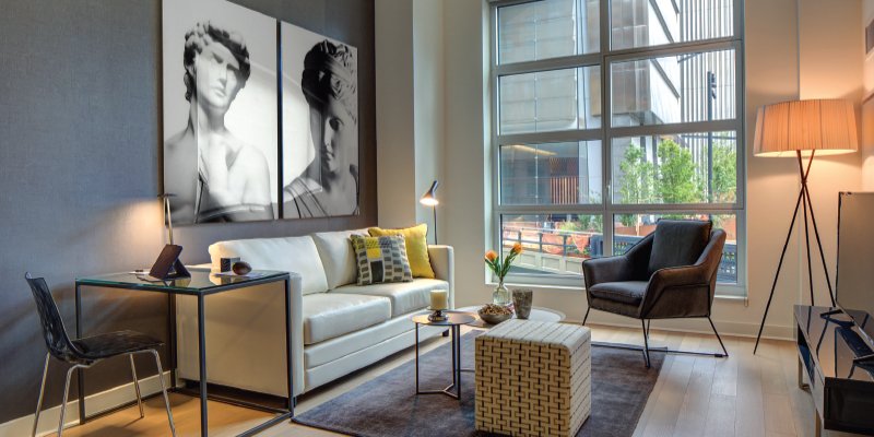 A modern, cozy living room with a sleek design. The space features a large window offering a view of a cityscape with greenery. Inside, there's a white sofa with decorative pillows, a glass-top desk with a black chair, and a stylish armchair near a standing lamp. A small coffee table and ottoman rest on a dark area rug. The wall displays black-and-white artwork depicting classical sculptures, adding an artistic touch to the sophisticated and inviting interior.