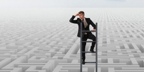 A businessman in a suit stands on a ladder above a vast, intricate maze, holding his hand over his eyes as he looks into the distance, symbolizing strategy, navigation, and problem-solving. The maze stretches out infinitely, blending into the horizon under a pale sky.