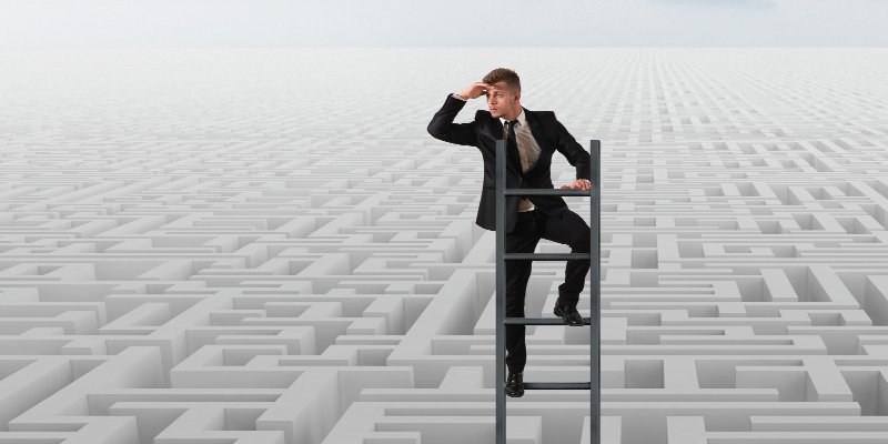 A businessman in a suit stands on a ladder above a vast, intricate maze, holding his hand over his eyes as he looks into the distance, symbolizing strategy, navigation, and problem-solving. The maze stretches out infinitely, blending into the horizon under a pale sky.