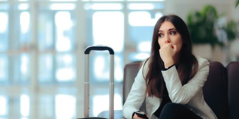 A young woman sits in an airport lounge with a suitcase beside her, looking pensive and anxious. She rests her hand near her mouth while holding her phone, appearing to be deep in thought. The blurred background features bright lighting and a soft-focus view of the terminal, emphasizing her contemplative mood.