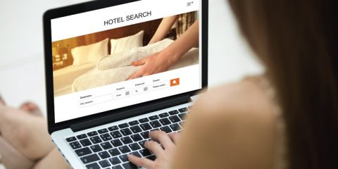 A person sitting indoors is using a laptop to browse a hotel booking website. The screen displays a clean interface with the words "HOTEL SEARCH" at the top and an image of a neatly folded towel being placed on a bed. Below the image is a form to enter details like destination, check-in and check-out dates, number of guests, and other preferences. The website design is modern and user-friendly, suggesting an online booking platform. The person is seated comfortably, emphasizing convenience and accessibility in travel planning.