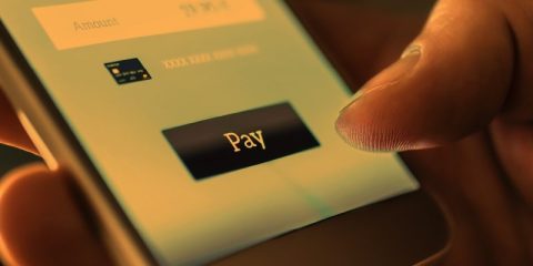 A close-up image of a person using a smartphone to make a payment. The screen displays a digital payment interface with a "Pay" button prominently featured, along with a partially visible credit card icon and amount field. The warm lighting highlights the fingertip pressing the button, symbolizing seamless mobile transactions and digital financial convenience.