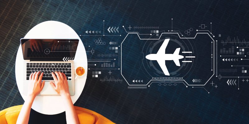 A top-down view of hands typing on a laptop placed on a white round table with an orange chair. The background features a futuristic digital overlay with a graphic of an airplane, surrounded by abstract technological symbols and interface elements. The image suggests themes of online travel booking, aviation technology, and modern connectivity.
