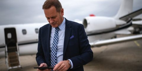 A businessman in a dark navy suit and a checkered tie is standing outdoors on an airport tarmac in front of a private jet. He is looking down and interacting with his smartphone, appearing focused and engaged. The private jet is white with a sleek design, its door open in the background, emphasizing luxury and executive travel. The overcast sky adds a neutral tone to the environment.