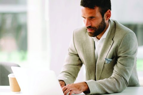 A well-dressed man in a beige suit works intently on a laptop in a bright, modern office setting. A cup of coffee sits beside him on the desk, emphasizing focus and productivity in a professional environment. The blurred background suggests a clean and open workspace.