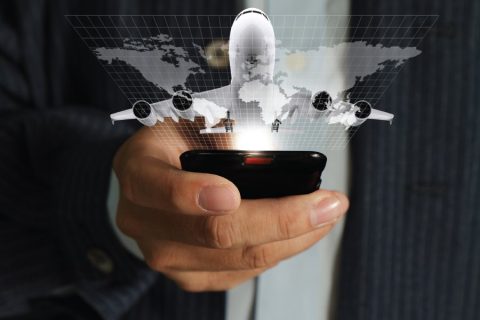 A businessman holding a smartphone projects a holographic image of an airplane flying over a world map. The grid overlay and bright light emanating from the phone emphasize modern technology and global connectivity. The image symbolizes advancements in mobile travel booking, international logistics, or the aviation industry's digital transformation.