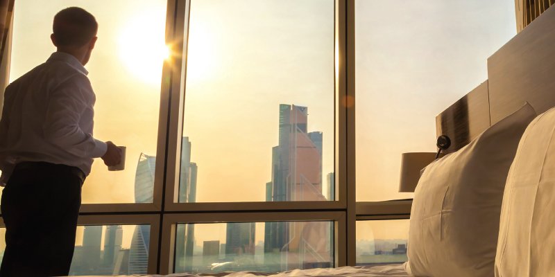 A business professional stands near a large window in a modern hotel room, holding a cup of coffee. The view outside showcases a cityscape with tall skyscrapers bathed in golden sunlight during sunrise or sunset, creating a serene and reflective atmosphere. The bed in the foreground adds to the luxury and comfort of the scene.