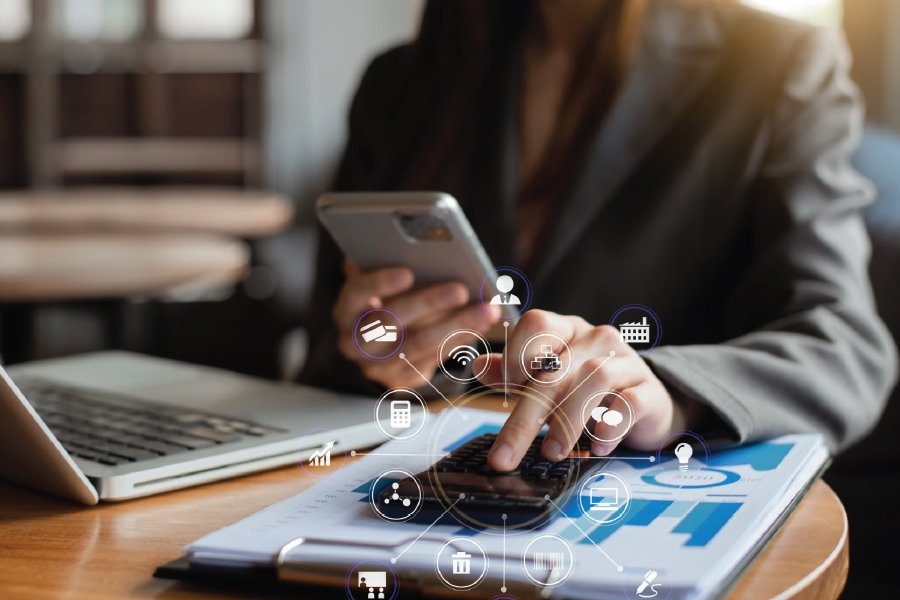 A person in a business suit is using a smartphone with one hand and a calculator with the other, sitting at a desk with a laptop and printed financial documents featuring bar graphs and charts. Digital icons representing various business and technology concepts (like a calendar, cloud storage, and communication tools) are overlaid on the image, symbolizing digital business solutions.