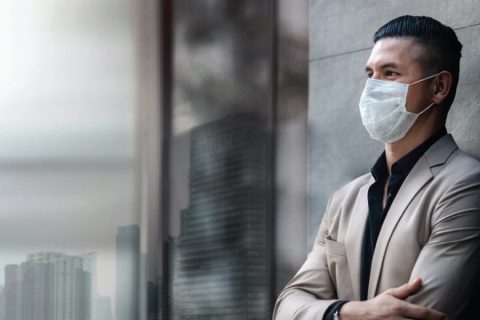 A confident man in a beige blazer and black shirt wearing a face mask stands with arms crossed against a modern building backdrop. The blurred cityscape visible through glass windows suggests an urban setting. The image reflects themes of professionalism, resilience, and health-consciousness in contemporary environments.