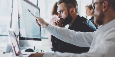 Two professionals are working collaboratively at a modern office. One, with glasses, is pointing at a computer screen displaying charts or data, while the other intently observes, deep in thought. Their focused expressions suggest strategic planning or data analysis. The background is softly blurred, showing other colleagues and a bright, open office environment.