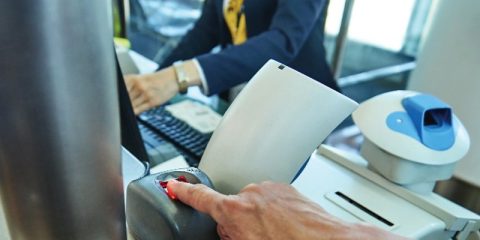 A close-up of a person's hand placing their fingertip on a fingerprint scanner at an airport check-in counter. The background shows a staff member working on a computer, dressed in a professional uniform with a yellow scarf. The setting highlights the integration of biometric technology in modern travel, ensuring security and efficiency in passenger identification.