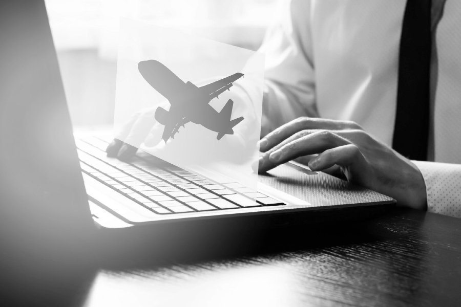 A monochromatic image of a person in formal attire typing on a laptop, with a transparent overlay of an airplane silhouette hovering above the keyboard. The image symbolizes online flight booking, digital travel management, or aviation-related tasks. The black-and-white tone emphasizes professionalism and focus.