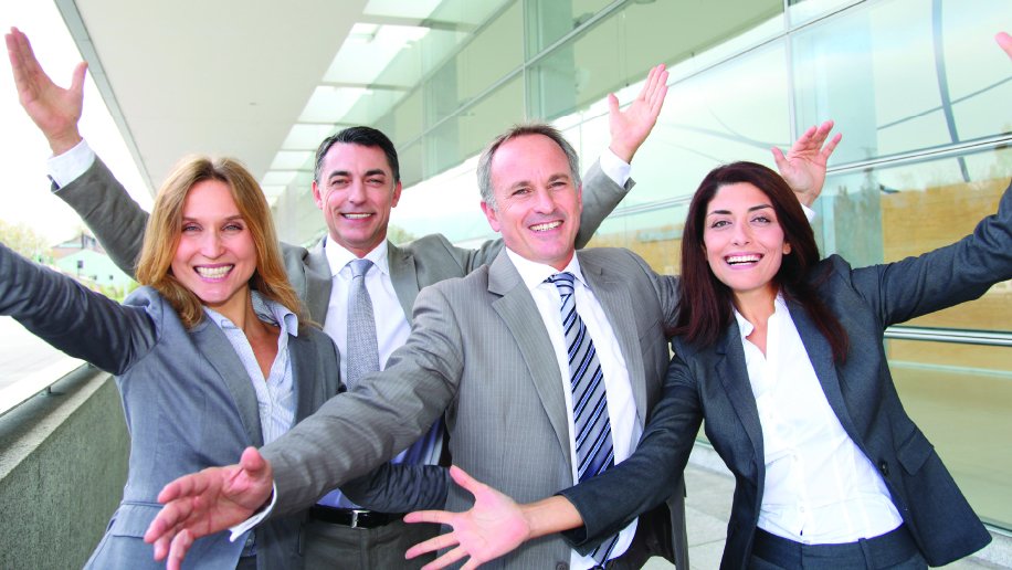 A group of four cheerful business professionals in suits, standing outside a modern office building with glass walls. They are smiling broadly with their arms raised in a celebratory gesture, symbolizing teamwork, success, and positivity in a corporate environment. The natural light and contemporary setting enhance the image's vibrant and optimistic tone.